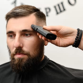 Barber shaves the temple with a cordless trimmer during a short haircut on the sides of the head.