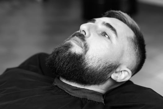 A young man with a beard in a black cape sits on a chair in a barbershop. The client is waiting for a haircut and shaving of the beard.