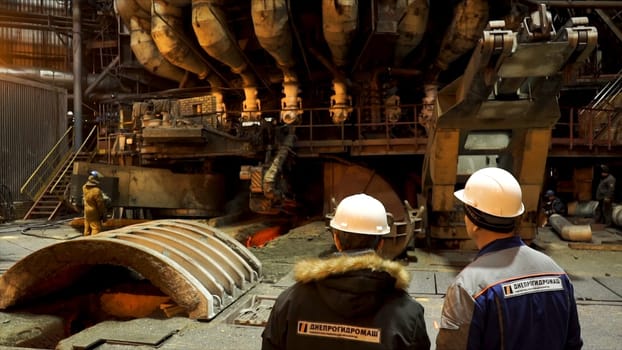 Rear view of two male workers in winter uniforms and white safety helmets moving forward to blast furnace, heavy metallurgy concept. Foundry in metallurgical plant, dark workshop with professional equipment.
