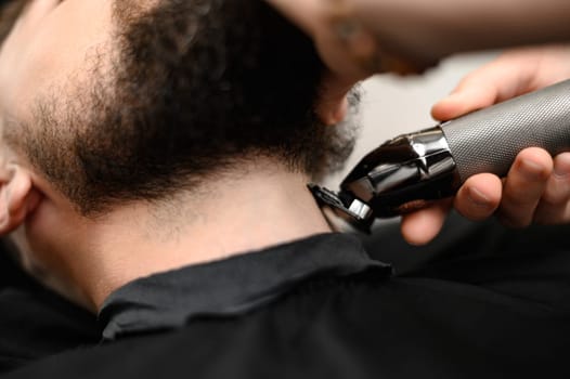 A barber in the salon trims the beard of a Caucasian man with a trimmer in the neck area