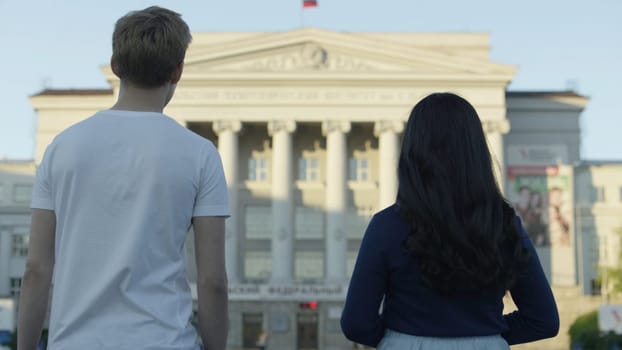Rear view portrait of two students carrying walking and look at the building. Back view of a couple of students sightseeing in a travel destination with a university with colorful buildings in the background