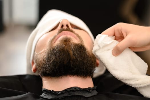 Hairdresser stylist wipes the remains of shaving gel with a towel after shaving the beard