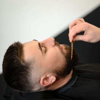 Barber combs beard with comb while cutting and shaving Caucasian man