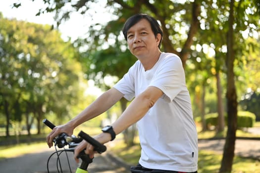 Portrait of happy retired man riding electric bicycle in public park on sunny day. Healthy lifestyle concept