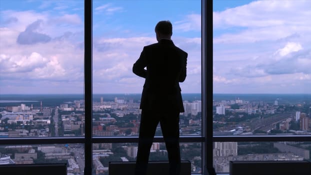 Businessman in suit standing next to a big window from an office building and checking his watch back view. Businessman standing at window looking on his wrist watch.
