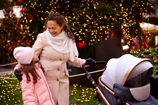 Happy smiling young woman, loving mother looking at her daughter, pushing baby pram, enjoy a Christmas atmosphere outdoors, walking along the beautiful street illuminated with garlands and lightings