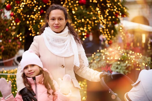 Beautiful mother with baby pram and her cute daughter looking at camera, standing against a Christmas tree outdoors, enjoying festive atmosphere during traditional family market. Bokeh on foreground