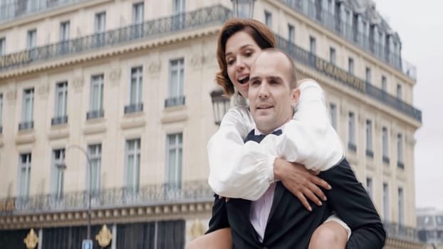 Portrait of a groom carrying happy bride on his back in the city street. Action. Husband and wife newlyweds at the video shooting outdoors