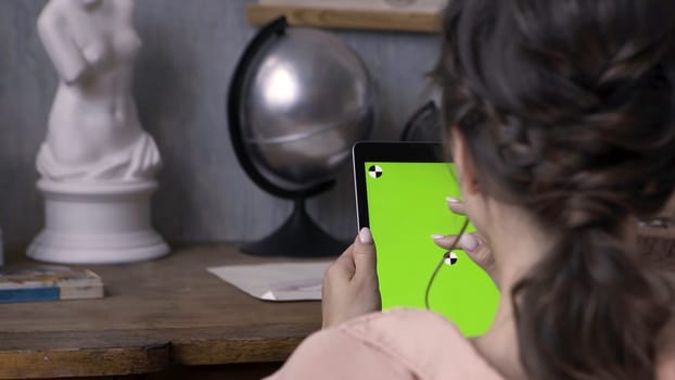 Woman hands holding tablet pc with green screen in the art studio with concrete wall and silver globe, plaster woman sculpture on the wooden table. Girl tapping on chromakey tablet.