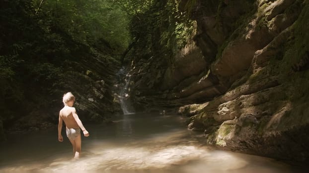 Boy goes into water in forest pond. Creative. Boy goes swimming in pond in rocks of forest. Forest pond with boy on sunny summer day.