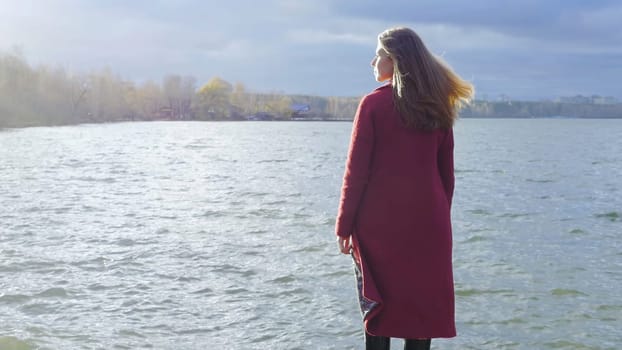 Young girl with her hair stands near the water in the cold season. Back view. Young woman standing near a lake.