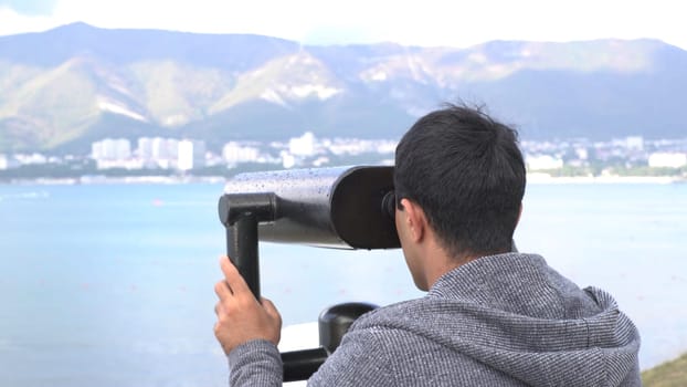 A man looking through coin binoculars on fantastic mountain and sea view. Male tourist looks at the coastal city through coin binoculars.