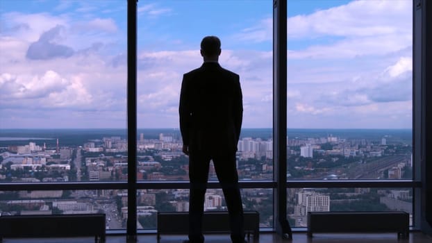 Businessman in suit standing next to a big window from an office building and checking his watch back view. Businessman standing at window looking on his wrist watch.