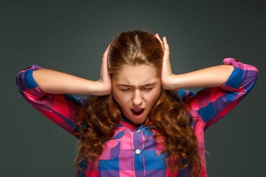 Young brunette woman with wavy hair, hands covering her ears, isolated on dark background.