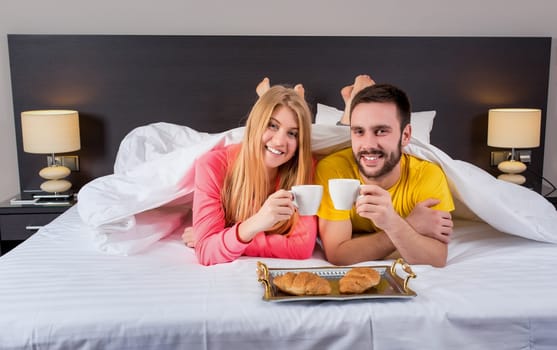 Happy young couple having breakfast tray on bed at home in bedroom. Couple drink coffee in bed, happy smile