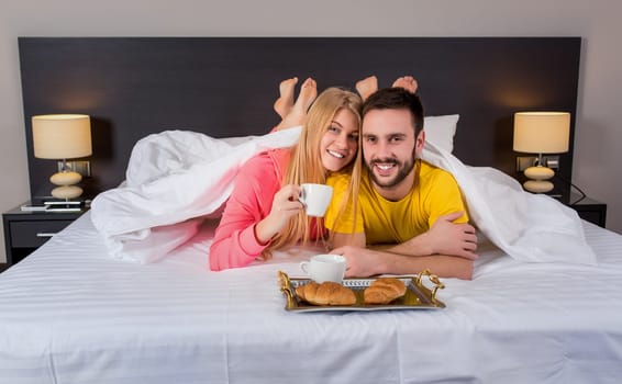 Happy young couple having breakfast tray on bed at home in bedroom. Couple drink coffee in bed, happy smile