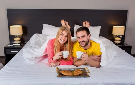 Happy young couple having breakfast tray on bed at home in bedroom. Couple drink coffee in bed, happy smile