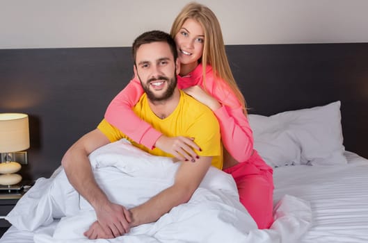 Young lovely couple have fun in a bed. Woman hugging man, happy smiles looking at camera