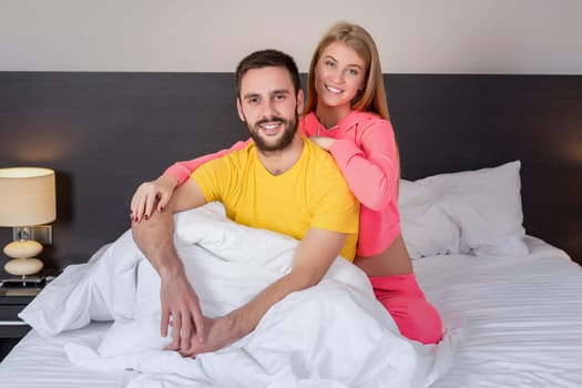 Young lovely couple have fun in a bed. Woman hugging man, happy smiles looking at camera