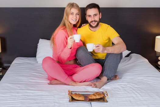 Happy young couple having breakfast tray on bed at home in bedroom. Young couple drink coffee in bed, happy smile