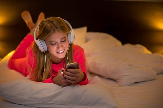 Young pretty blond women smiling listening to the music with a headset from a smartphone lying on the bed at home