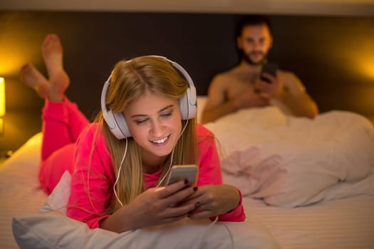 Young happy women on white bed using mobile phone with headphones, boyfriend on background. Concept about technology and people