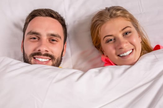 Young lovely couple lying in a bed, happy smile looking at camera, cover under blanket top angle view up above