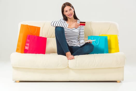 Online shopping and technology concept - smiling beautiful young woman with tablet pc computer and credit card. She sit on sofa with colorful bags