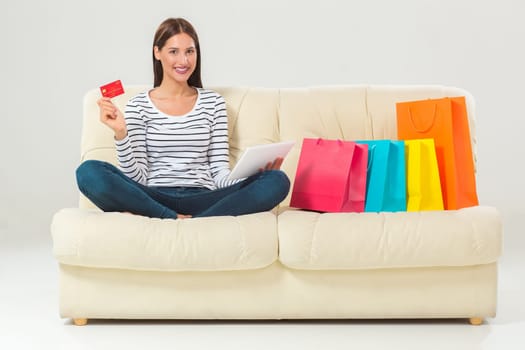 Online shopping and technology concept - smiling beautiful young woman with tablet pc computer and credit card. She sit on sofa with colorful bags