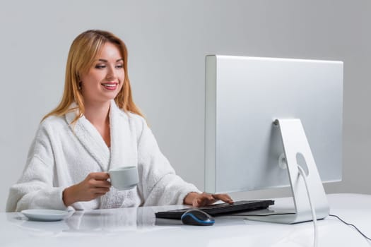 young woman sitting in the table and using computer on gray background. girl smiling and looking at monitor. with a cup in hands