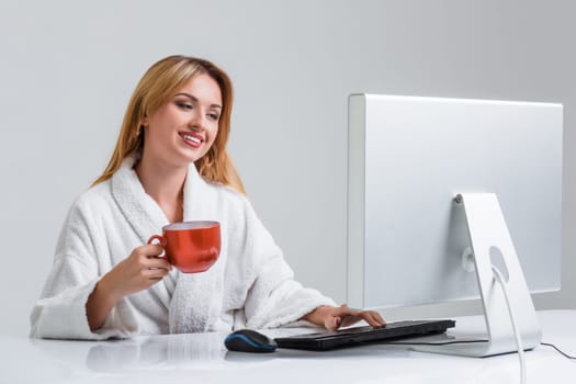 young woman sitting in the table and using computer on gray background. girl smiling and looking at monitor. with a cup in hands