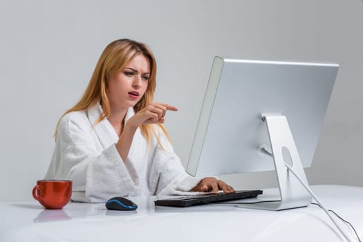 young woman sitting in the table and using computer on gray background. indignation is pointing at the monitor