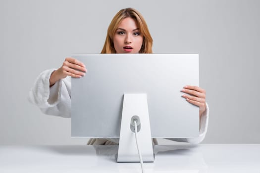 young woman sitting in the table and using computer on gray background. Surprised and scared hidden behind the monitor. It peeping from behind him