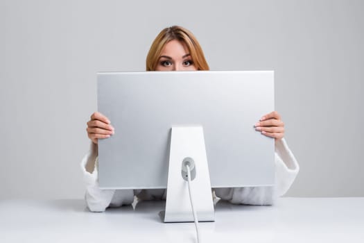 young woman sitting in the table and using computer on gray background. Surprised and scared hidden behind the monitor. It peeping from behind him