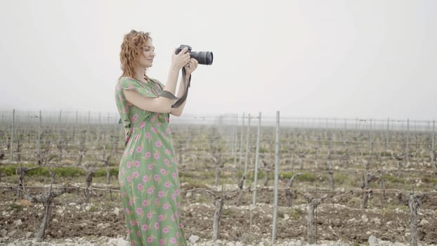 Young pretty woman takes pictures in harvest field. Action. Side view of female photographer in a field on grey sky background