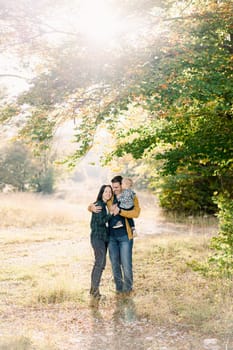 Dad hugging mom holding little girl in arms in sunny park. High quality photo