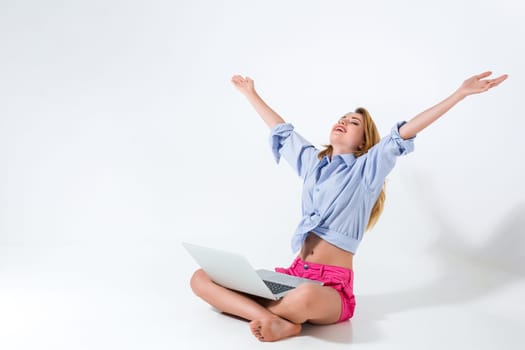 young woman sitting on the floor with crossed legs and using laptop on white background. very satisfied, happy, she raised her hands up