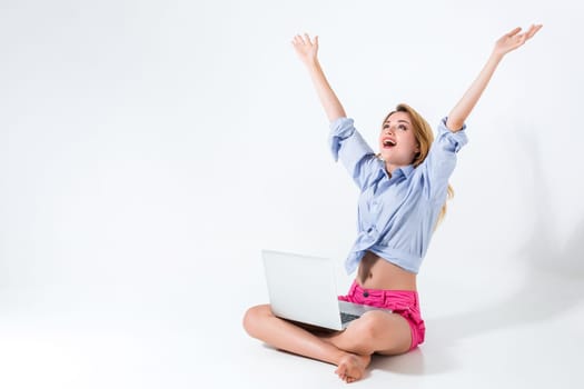 young woman sitting on the floor with crossed legs and using laptop on white background. very satisfied, happy, she raised her hands up