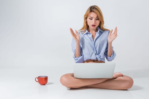 young woman sitting on the floor with crossed legs and using laptop on white background. Surprised, emotional