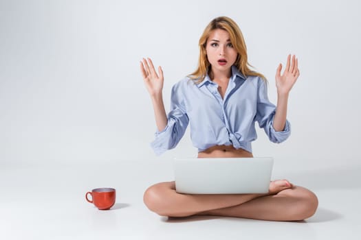 young woman sitting on the floor with crossed legs and using laptop on white background. Surprised, emotional
