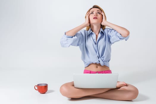 young woman sitting on the floor with crossed legs and using laptop on white background. tired, surprised, tortured, his hands holding his head