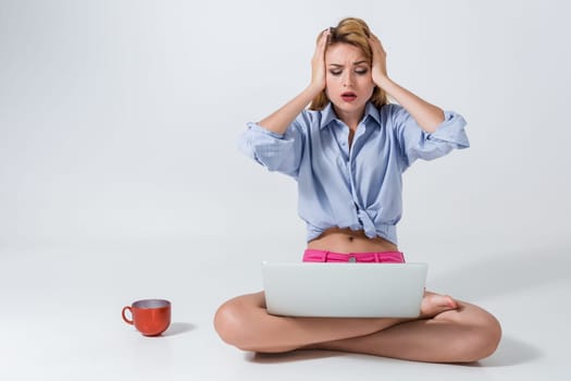 young woman sitting on the floor with crossed legs and using laptop on white background. tired, surprised, tortured, his hands holding his head