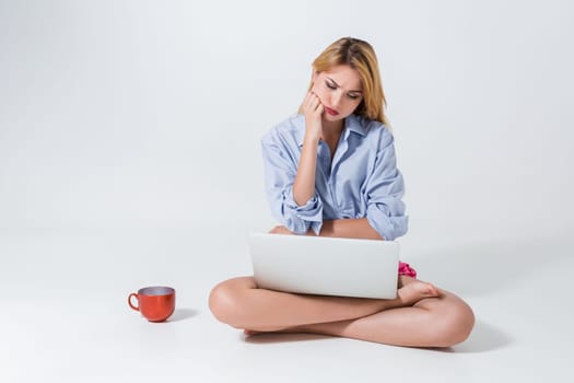 young woman sitting on the floor with crossed legs and using laptop on white background. sad, thoughtful look in laptop