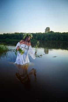 Adult mature brunette woman in a white dress, sundress and a wreath of flowers in summer in water of river or lake in evening at sunset. Celebration of the Slavic pagan holiday of Ivan Kupala