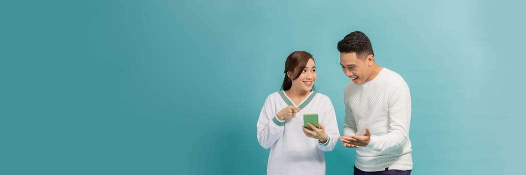 Amazed attractive young Asian couple looking at smartphone together isolated on blue background