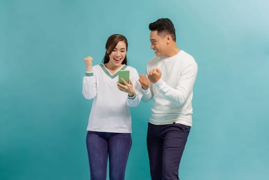 Amazed attractive young Asian couple looking at smartphone together isolated on blue background