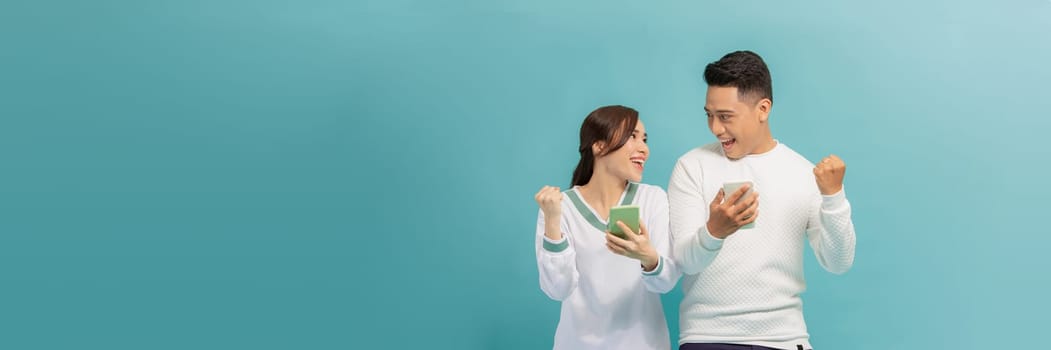 Cheerful attractive couple standing isolated over blue, using mobile phone, celebrating success