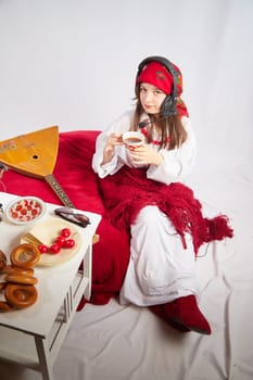 A girl in traditional national russian dress with a scarf and shawl, with musical balalaika and modern headphones with a microphone and glasses at a set table for Easter and Maslenitsa