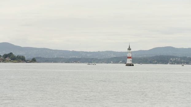Picturesque natural landscape with a lighthouse and rippling water surface. Action. Cloudy sky above ocean and mountains on the background