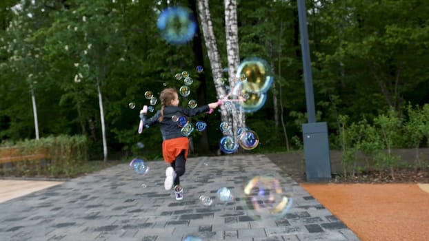 Lovely girl blowing bubbles while running, rear view. Female caucasian child dressed in warm clothes creating many soap bubbles in spring time on the background of green trees of a city park.